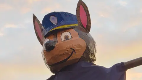 Amber Cummins A smiling paper mache German Shepherd dog with a police cap perched on its head. The figure has arms spread wide and is wearing a blue sheet. 