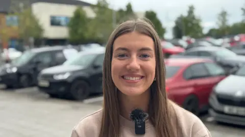 BBC Aliyah Rafferty outside her school, she is smiling wearing a beige jumper with long brunette hair. 

Behind her is a car park.  