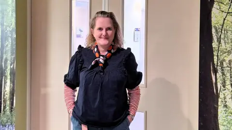 Jo Argyle is standing in front of a hospital door with her hands in her jeans pockets. She is smiling at the camera.