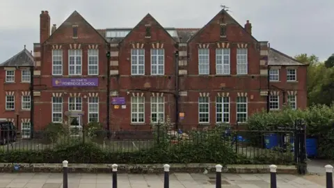 A google view of the front of Penbridge Junior School. It is a large red brick building with several white-framed Georgian windows. The school's gates are in the foreground which are lined by hedges.