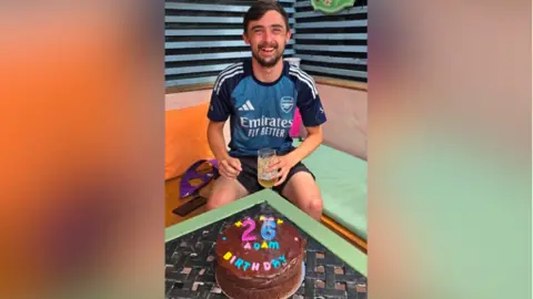 Family handout An image of smiling Adam McClelland with a birthday cake in front of him, in celebration of his 26th birthday. He is holding a glass with a drink. He is sitting in what appears to be an outdoor area.