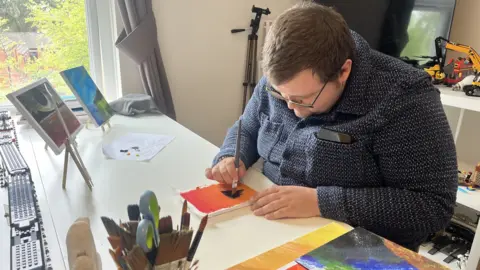 A man with brown hair, brown facial stubble and black glasses wearing a blue and white long sleeved top. He is leaning over a white table, painting on a small canvas. The painting has an orange and red ombre background and he is beginning to paint a black floating island. There are other canvases and painting supplies around him