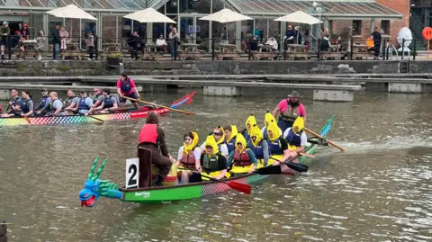 Two boats with 10 paddlers each are on the water in a city's docks, paddling towards the camera
