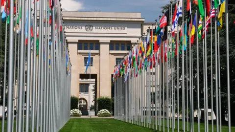 Reuters Two rows of national flags on metal poles line each side of a green lawn. They are in front of a large stone building that has the words 'Nations Unite' on the front.