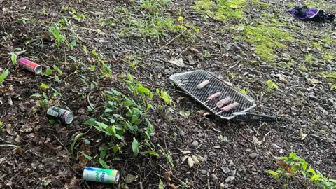 Russell Millner Uncooked sausages and kebabs left on a barbeque on a patch of outdoor ground. There are alcoholic cans discarded too.
