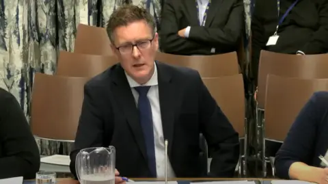 NI Assembly  Dr Andrew Brown, sitting in the education committee. He has spikey brown hair and wide glasses and wears a suit. 