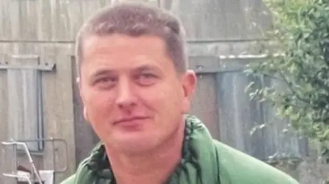 A smiling Paul Ainscough, with short cropped brown hair and wearing a green jacket, stands in front of a grey concrete wall and a green bush