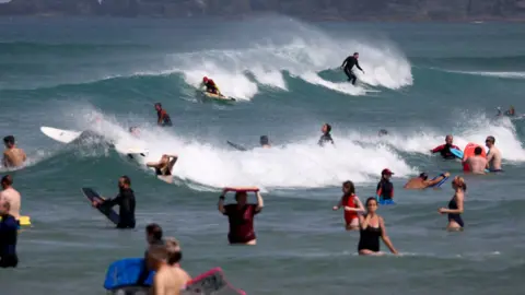 A number of surfers and swimmers in the sea, some are holding bodyboards and some are surfing.