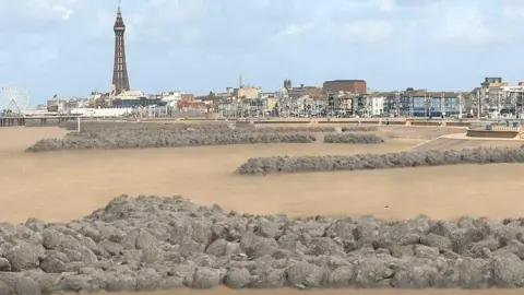 Visualisation of rock headlands along the beach. They are long stretches of grey rocks edging from the steps to the beach towards the sea. Blackpool's coastline can be seen in the background, with the tower to the left.