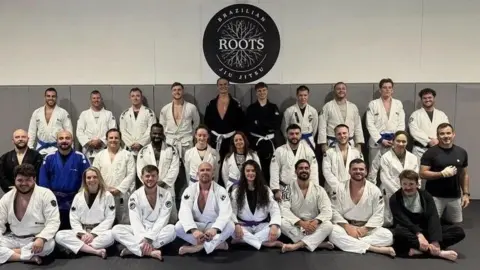 Bea Fernandes/Roots Gym A series of Jiu-Jitsu fighters pose infront of the Roots Gym logo dressed in white combat attire.
