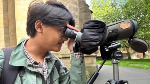 BBC/Simon Thake An man with dark floppy hair peers through the lens of a covered device like a telescope