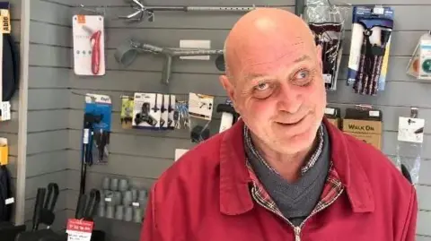 A man smiles at the camera. He is wearing a red jacket and is standing in the new Easy Mobility services showroom. Behind him are various mobility aids.
