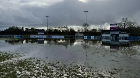 The pitch seen from ground level with large puddles of water still visible and a small grandstand with white seats in the background