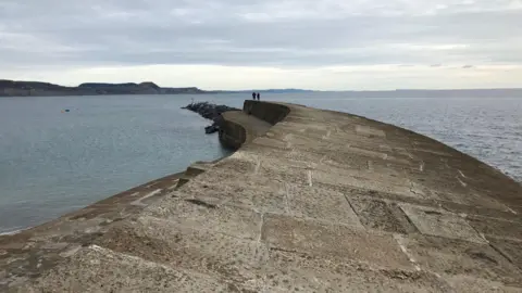 BBC The top of the cobb as it runs into the sea. There are people at the end of it going for a walk.