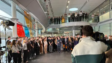 Sir Kier Starmer addresses staff at the McLaren Group headquarters in Woking. In the foreground, we can see the back of a camera person who is filing the event.