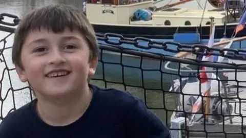 Family picture Robin Caliskan with short brown hair smiling wearing a long sleeve navy top with different signs on it. Behind he is standing by a chained gate with boats and a canal in the background.