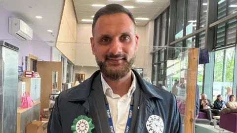 BBC/Aisha Iqbal Sajawal standing indoors in a bright, modern public building. He is positioned centrally in the foreground, facing the camera directly. He has short hair and a neatly trimmed beard and is wearing a dark jacket over a white collared shirt. Around the neck, there is a blue lanyard with the word “STAFF” printed repeatedly in white capital letters.
Attached to the front of the jacket are several badges and symbols. A green rosette is pinned to one side of the chest, and there are additional circular badges and a small ribbon pin nearby.
