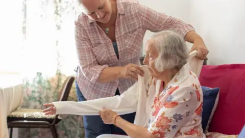 Getty Images Carer helps elderly lady put on her jacket. Carer is standing over the woman who wears a colourful flowery blouse and has white hair