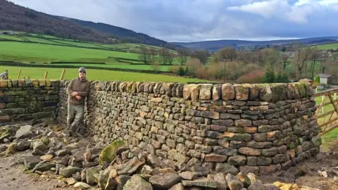 Phil Dolphin A man wearing a light brown hoodie, grey jeans, sunglasses and a grey woolly hat leans against a dry stone wall, which is about the same height as he is. 