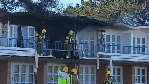 Firefighters are pictured tackling a blaze at an upper-storey beach hut. Two of them can be seen up on a balcony, where part of the building is blackened and badly damaged, while others look on from the ground. 