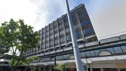 A five-storey office block viewed from an angle slightly to the side rising above a shopping centre. It has brown bricks and rectangular windows that are all the same size.