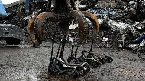 Northamptonshire Police A row of e-scooters that are about to be crushed by a large metal claw. Metal items are piled up behind. 