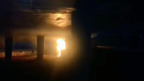 A picture taken of the fire. The flames from the campervan have reached the underside of the bridge. Smoke is spreading out under the bridge from the campervan.