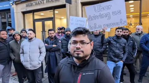 Mohammed Rahman poses for the camera while his colleagues stand behind him holding signs to protest against pay at Uber.