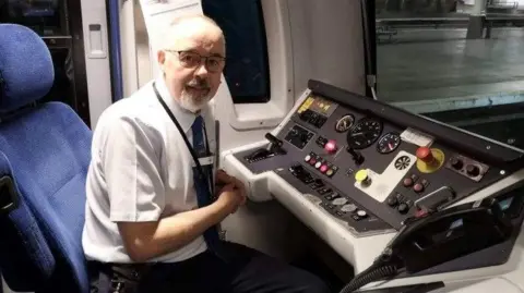 TPE Don Coffey sat in the train driver cabin with a dashboard in front of him. He is wearing a white short-sleeved shirt and dark tie.