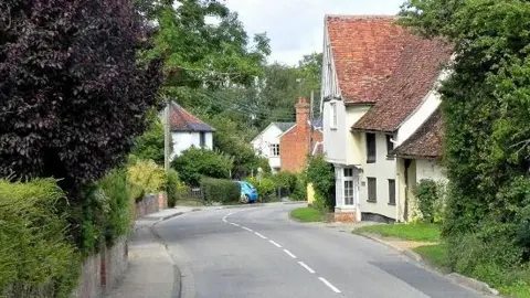 The Street in Monks Eleigh. The road is lined by small cottages, tall trees and leafy bushes. It is a 30 miles per hour limit and is slightly bendy.