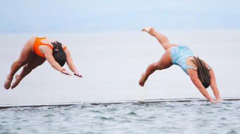 PA Media is wearing two brunette women - an orange swims suit on the left and the woman on the right is wearing a light blue and white printed swims suit - Diving in Clevedon Marine Lake in Clevedon.