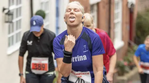 Bridgnorth Running Club A female runner signals her tiredness on a steep hill by pulling a face with her tongue hanging out. The woman has blonde hair and is wearing a purple t-shirt, sweatbands on her wrists and earphones. A running number is just visible pinned to her t-shirt, saying Bridgnorth 10k. Other female runners are visible out of focus behind her.