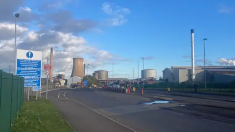 Jo Makel/BBC A view of Scunthorpe steelworks. A wide road runs towards cooling towers, metal chimneys and large buildings. To the left is a green fence and a large sign with white text on a blue background.