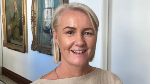 Tracy Kelly is standing in a white hallway with two large painted portraits in the background. Kelly has short, blonde hair and she is smiling at the camera. She is wearing a beige jumper.