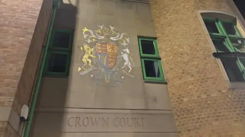 Brian Farmer/BBC Luton Crown Court: The words "CROWN COURT" are written below a multi-coloured crest on a grey wall above an entrance to a brown-brick building with green-framed windows.