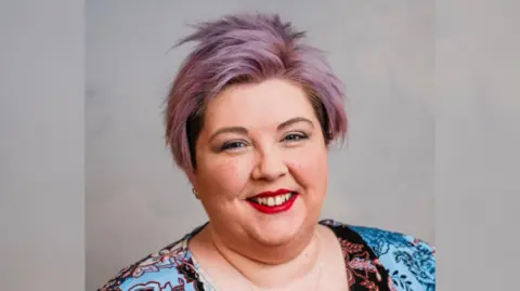 Ciara Hunter Ciara Hunter has short purple hair and is wearing makeup with red lipstick. She has a multicoloured top which is blue and black. The background is grey.