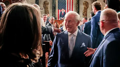King Charles, with a grin on his face, walks by lawmakers in US Congress after delivering his speech, with US Vice-President JD Vance and a US flag visible distantly behind him