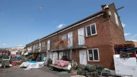 Two-storey red-brick buildings with iron stairs accessing the second floor. They have white doors and windows, with fishing industry equipment such as nets strewn outside the building.