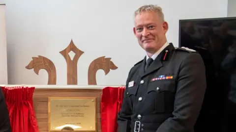 East Sussex Fire and Rescue Service A man in a fire officer's uniform stands smiling next to a commemorative plaque which is framed by red curtains