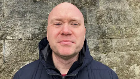 Declan Campbell is bald and clean shaven and is wearing a navy coat with a high collar