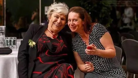 Ann Jessopp and Sarah Johnson. Ann has short white hair and is wearing a black and red dress with a black jacket. Sarah is sat next to her. She has long brown hair and is wearing a black and white spotted dress. They are both looking at the camera and smiling. 