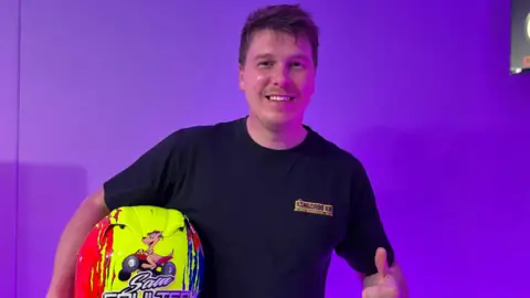 BBC A man posing with his left thumb up against a purple-lit backdrop, holding a red and yellow helmet in his right hand with a picture of a kangaroo on a quad bike. He is wearing a black t-shirt saying Kangaroo kid. He has brown hair and is smiling.