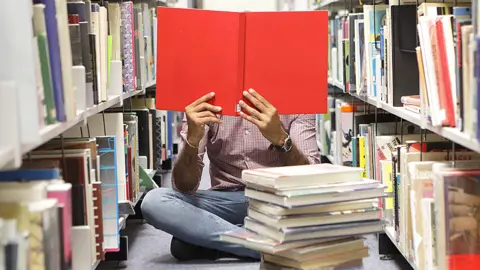 Person sitting on the floor holding up an open book covering their face while surrounded by shelves of books and books piled up on the carpet in front of them