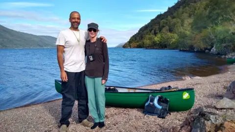 Family Handout Tarig Sinada and Jenny Cawson. The couple are standing next to each other near a lake. A green canoe is on the shore