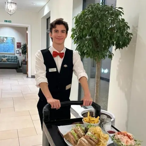 University of Essex Fernando is dressed in a white shirt and black waistcoat. He has a red bow tie. He is pushing a black trolley with food on it.