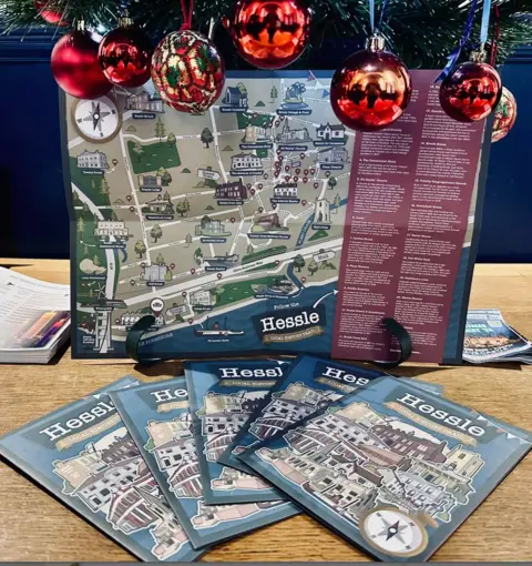 Hessle Local History Society Picture of the opened-out map with notes of points of interest on the right hand side and five booklets of the map in front of it. Christmas baubles dangle about the foled-out map.