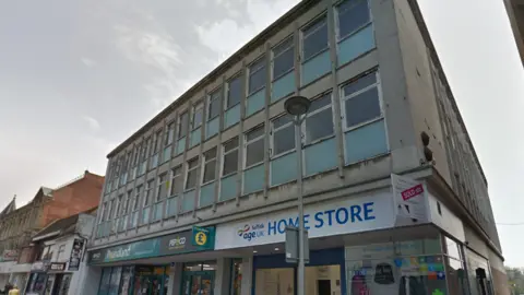 Carr Street (pictured in 2019)  has a row of shops and above a two storey building with windows. Beside it are several more adjoining buildings with roofs at different levels and angles.