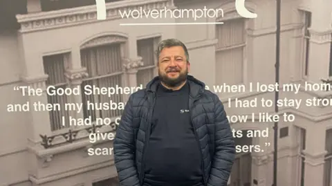 Jack is stood in front of a poster for the charity. He has a black jacket and jumper on. He has a beard and greying hair.