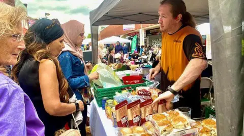 A man with long hair tied back at a food stall which has what looks like scones and packets on it. Three women are looking at the products on sale. 