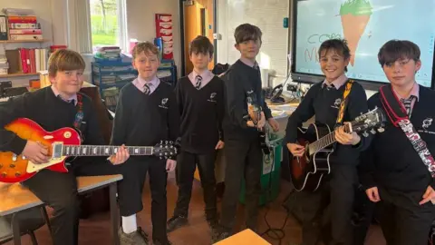 A group of young lads all in school uniforms, in a classroom. Four of them have guitars and all look and smile at the camera.